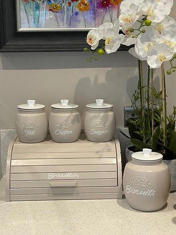 Ceramic jars labeled Tea, Coffee, and Sugar on a counter with a bread bin labeled Breadbin. White orchids and a colorful painting in the background.